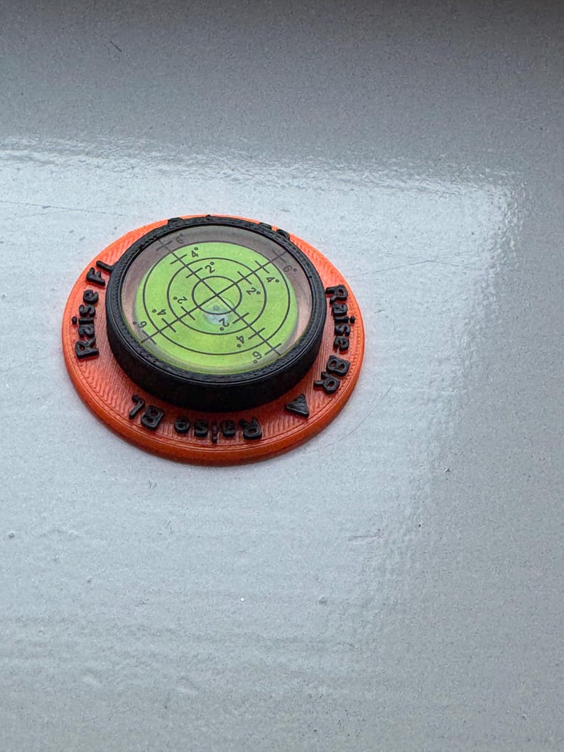 May include: A circular orange 3D-printed base with a black rim and a central green bullseye spirit level. The orange base features black embossed text reading Raise FL, Raise BR, and Raise BL with directional arrows for levelling adjustments.