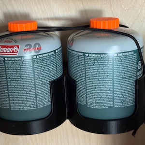 May include: Two Coleman propane canisters with orange lids, held in a black bracket. The tanks are labelled with text including "PROPANE" and "BUTAN/PROPAN BLANDING". A black cord connects the canisters.