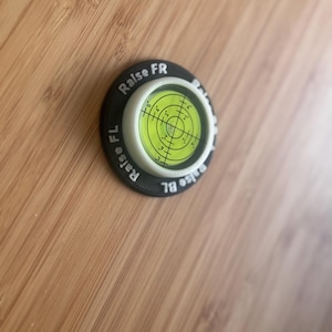 May include: A circular bullseye spirit level with a bright yellow-green liquid centre and black crosshair markings. The black outer ring features white printed text reading Raise FR, Raise FL, and Raise BL, indicating adjustment points for levelling.