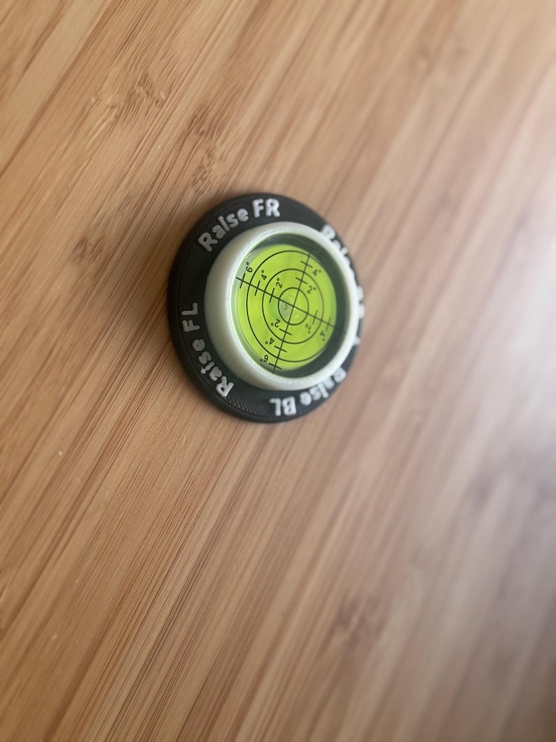 May include: A circular bullseye spirit level with a bright yellow-green liquid centre and black crosshair markings. The black outer ring features white printed text reading Raise FR, Raise FL, and Raise BL, indicating adjustment points for levelling.
