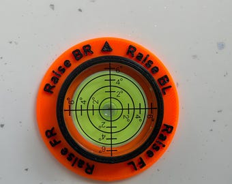 May include: A circular orange levelling tool with a central green liquid-filled bubble vial featuring black crosshair markings and degree indicators. The orange frame has black text printed around the perimeter reading Raise BR, Raise BL, Raise FR, and Raise FL.