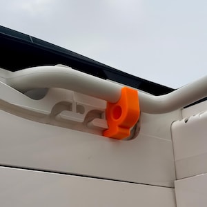 May include: A close-up shot of a white and black structure with a curved, light grey handle. An orange plastic piece is attached to the handle. The background is a cloudy sky.