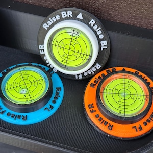 May include: Three circular bubble levels with bright yellow liquid centres and black crosshair markings. Each unit features a coloured base in black, blue, or orange, with embossed text reading Raise BR, Raise BL, Raise FR, and Raise FL around the rim.
