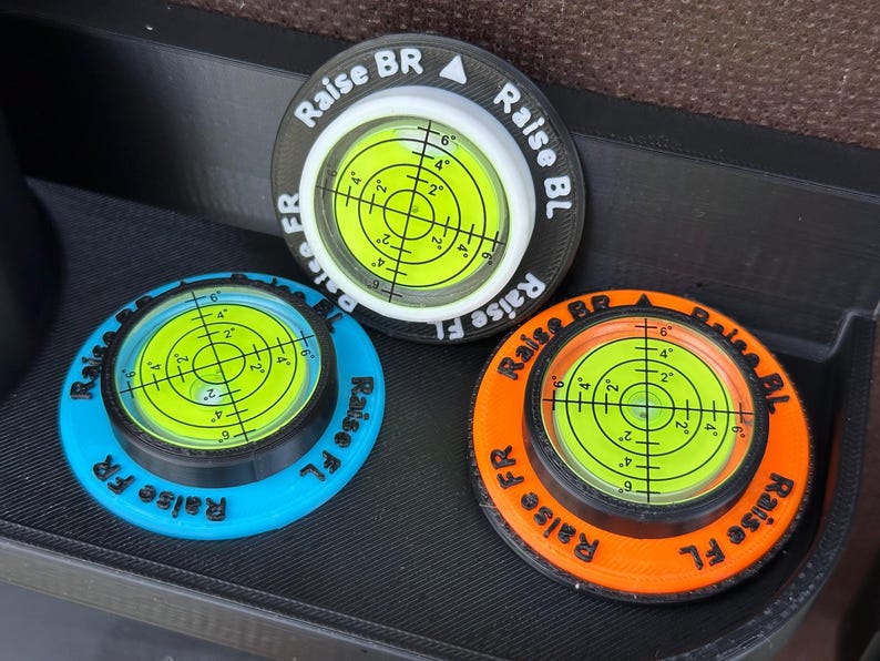 May include: Three circular bubble levels with bright yellow liquid centres and black crosshair markings. Each unit features a coloured base in black, blue, or orange, with embossed text reading Raise BR, Raise BL, Raise FR, and Raise FL around the rim.