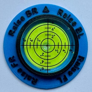 A circular motorhome or campervan level with a green bubble level and black markings. The level is surrounded by a blue ring with black text that reads 'Raise Fl', 'Raise FR', 'Raise BR A', and 'Raise BL'.