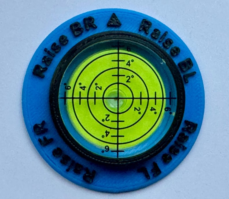 A circular motorhome or campervan level with a green bubble level and black markings. The level is surrounded by a blue ring with black text that reads 'Raise Fl', 'Raise FR', 'Raise BR A', and 'Raise BL'.
