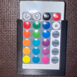 May include: A rectangular remote control in a black frame. It has buttons for on/off, colour selection (red, green, blue, white, and others), and light effects (flash, strobe, fade, smooth). The buttons are round and brightly coloured.