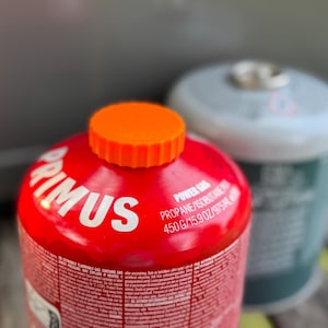 May include: Two gas canisters are visible. The main one is red with white text and an orange cap, branded "PRIMUS". The second is silver and green. The red canister contains 450g of propane/isobutane.