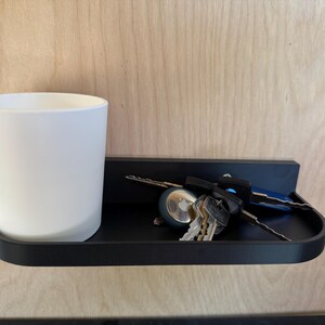 May include: A black wall-mounted shelf holding a white mug and a set of keys. The shelf has a curved front edge and is fixed to a light wood-grain surface. The keys include a car key and several house keys.