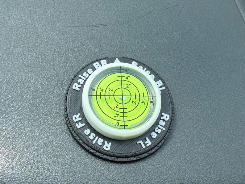 May include: A circular black levelling tool with a central lime green bullseye spirit level. The black surface features white text reading Raise BR, Raise BL, Raise FR, and Raise FL, with a crosshair grid marked with 2, 4, and 6 degree increments.