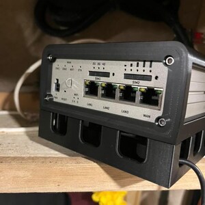 May include: A black and silver network device with multiple ports and indicator lights. The device has labels for LAN, WAN, and SIM slots. It is mounted in a black frame and sits on a wooden shelf. Cables are visible in the background.