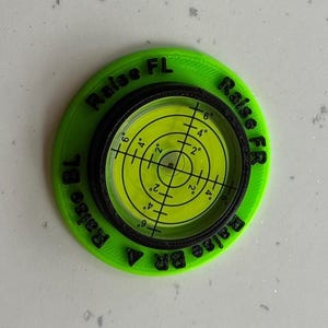 May include: A circular lime green levelling tool with a black centre containing a yellow liquid-filled bullseye level. The green base features black embossed text reading Raise FL, Raise FR, Raise BL, and Raise BR around the central gauge.