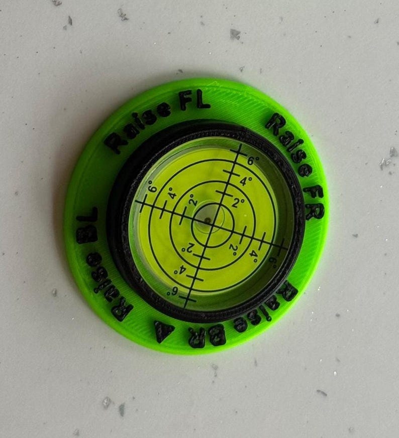 May include: A circular lime green levelling tool with a black centre containing a yellow liquid-filled bullseye level. The green base features black embossed text reading Raise FL, Raise FR, Raise BL, and Raise BR around the central gauge.