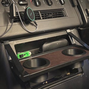 Op de afbeelding: Een zwarte, neerklapbare bekerhouder voor in de auto met twee ronde openingen. De bekerhouder heeft een bruin patroon en is gemonteerd in het dashboard van een voertuig. Een groen voorwerp en een zwart etui liggen in het open vak erboven.