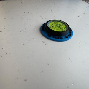 May include: A circular bullseye spirit level featuring a bright neon green liquid vial with black crosshair markings, encased in a black frame and mounted on a flat blue base with black text embossed along the outer edge of the blue circular frame.
