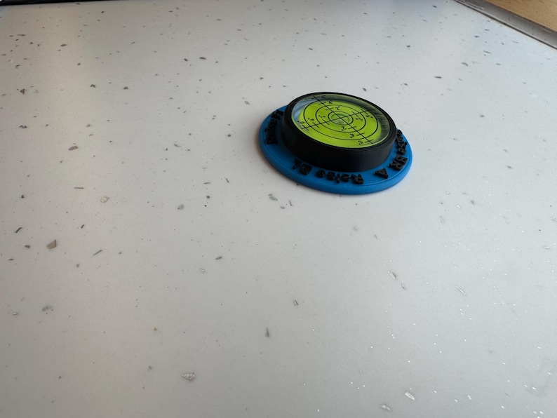 May include: A circular bullseye spirit level featuring a bright neon green liquid vial with black crosshair markings, encased in a black frame and mounted on a flat blue base with black text embossed along the outer edge of the blue circular frame.