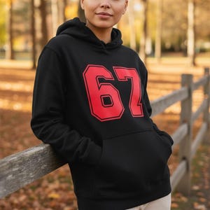 Op de afbeelding: Zwarte hoodie met het nummer 67 in rode blokletters. De sweatshirt heeft een voorzak en een capuchon met trekkoord. De persoon leunt tegen een houten hek in een buitenomgeving.