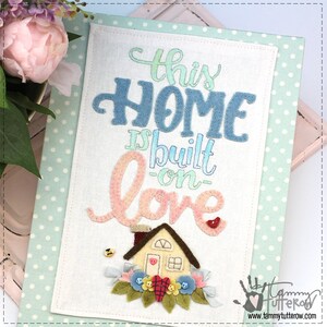 May include: A white felt wall hanging with the text "This Home is built on love" in colorful lettering. A small house with a red heart on the door is surrounded by flowers and leaves. The text "Tammy Tutterow" is in the bottom right corner.