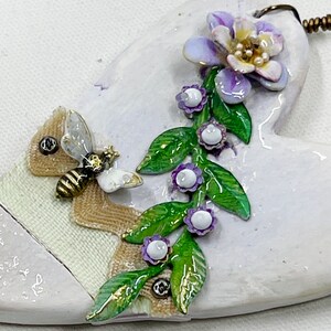 May include: A white ceramic heart-shaped ornament with a bee, a vine of purple flowers, and green leaves. The ornament has a gold-colored string for hanging.