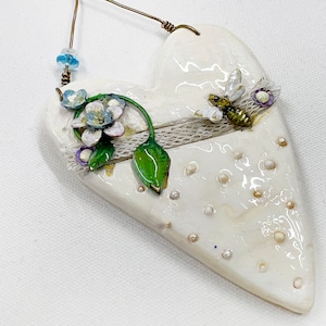 May include: White ceramic heart-shaped ornament with a floral design, a bee, and white beads. The ornament has a wire hanger.