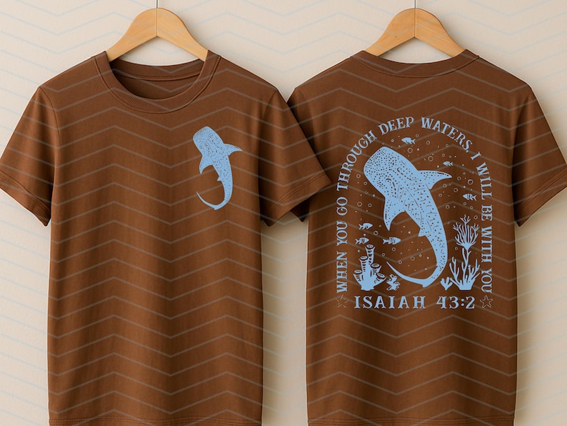 May include: Brown t-shirt with a light blue whale shark graphic on the front and back. The back of the shirt features the text "WHEN YOU GO THROUGH DEEP WATERS, I WILL BE WITH YOU" and "ISAIAH 43:2". The shirt has a chevron pattern.