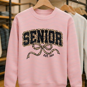 May include: A light pink sweatshirt with the word "SENIOR" in black block letters with a leopard print fill. Below the word is a leopard print bow with the text "EST. 2026". The sweatshirt is hanging on a wooden hanger.