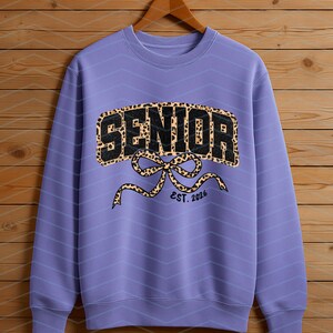 May include: A lilac-colored sweatshirt with a leopard print "SENIOR" design. The design includes a bow and the text "EST. 2026". The sweatshirt is hanging on a wooden hanger against a wooden background.