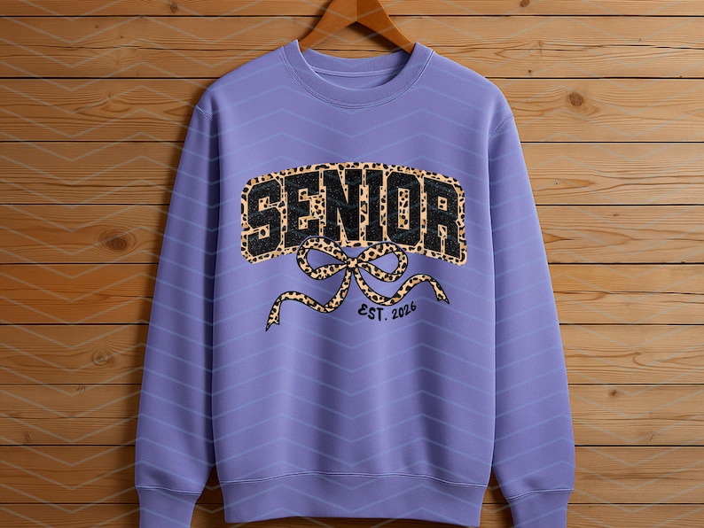 May include: A lilac-colored sweatshirt with a leopard print "SENIOR" design. The design includes a bow and the text "EST. 2026". The sweatshirt is hanging on a wooden hanger against a wooden background.