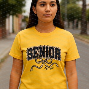 May include: A yellow t-shirt with the word "SENIOR" in black leopard print lettering. Below the word "SENIOR" is a bow with the text "EST. 2026". The shirt is short-sleeved.