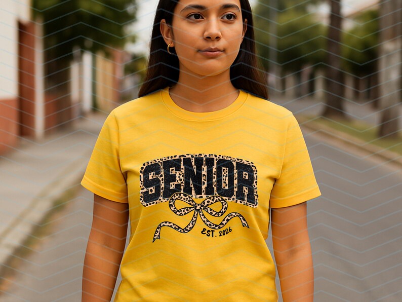 May include: A yellow t-shirt with the word "SENIOR" in black leopard print lettering. Below the word "SENIOR" is a bow with the text "EST. 2026". The shirt is short-sleeved.