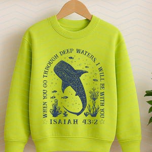 May include: A lime green sweatshirt featuring a blue whale shark design with the text "WHEN YOU GO THROUGH DEEP WATERS, I WILL BE WITH YOU. ISAIAH 43:2". The design includes fish and coral. The sweatshirt is hanging on a wooden hanger.