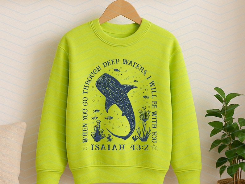 May include: A lime green sweatshirt featuring a blue whale shark design with the text "WHEN YOU GO THROUGH DEEP WATERS, I WILL BE WITH YOU. ISAIAH 43:2". The design includes fish and coral. The sweatshirt is hanging on a wooden hanger.