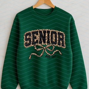 May include: A dark green sweatshirt with the word "SENIOR" in a leopard print design. Below the word is a leopard print bow and the text "EST. 2026". The sweatshirt is hanging on a wooden hanger.