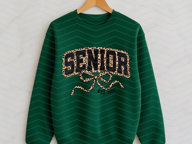 May include: A dark green sweatshirt with the word "SENIOR" in a leopard print design. Below the word is a leopard print bow and the text "EST. 2026". The sweatshirt is hanging on a wooden hanger.