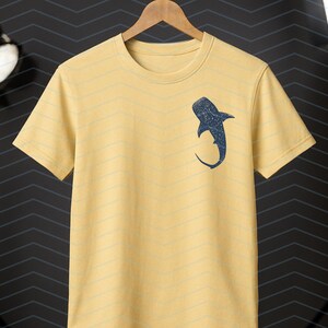 May include: Yellow short-sleeved t-shirt with a navy blue whale shark graphic on the front. The shirt is hanging on a wooden hanger against a dark background. The shirt has a crew neck and a relaxed fit.