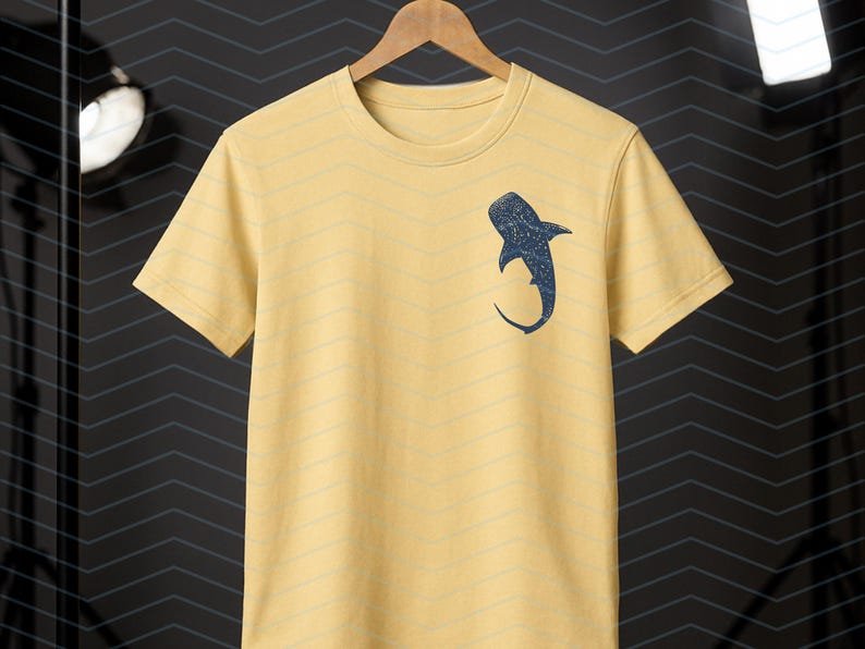 May include: Yellow short-sleeved t-shirt with a navy blue whale shark graphic on the front. The shirt is hanging on a wooden hanger against a dark background. The shirt has a crew neck and a relaxed fit.