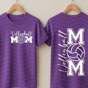 May include: A purple t-shirt featuring a white volleyball graphic and the text "Volleyball Mom". The back of the shirt displays the text "Volleyball Mom" with a volleyball and heart design. The t-shirt is displayed on a wooden hanger.