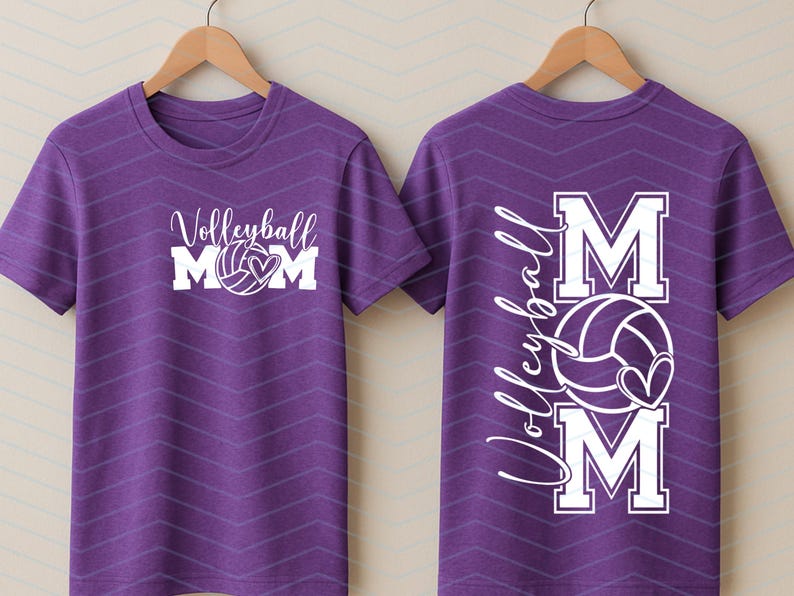 May include: A purple t-shirt featuring a white volleyball graphic and the text "Volleyball Mom". The back of the shirt displays the text "Volleyball Mom" with a volleyball and heart design. The t-shirt is displayed on a wooden hanger.