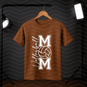 May include: A brown t-shirt with a white graphic. The design includes the word "MOM", a volleyball, and the word "Volleyball". The t-shirt is displayed on a wooden hanger. The background is dark.