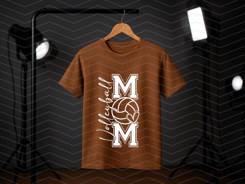 May include: A brown t-shirt with a white graphic. The design includes the word "MOM", a volleyball, and the word "Volleyball". The t-shirt is displayed on a wooden hanger. The background is dark.