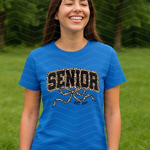 May include: A blue t-shirt with the word "SENIOR" in a leopard print design. Below the word "SENIOR" is a bow design with the text "EST. 2024". The shirt is worn by a person in a green outdoor setting.