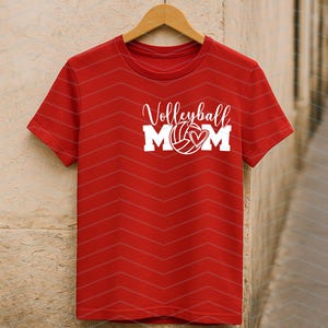 May include: Red t-shirt with the words "Volleyball MOM" in white script and block letters. A volleyball with a heart is in the center of the text. The shirt is hanging on a wooden hanger.
