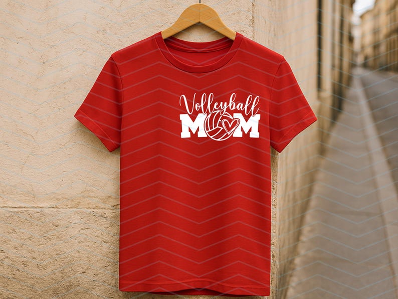 May include: Red t-shirt with the words "Volleyball MOM" in white script and block letters. A volleyball with a heart is in the center of the text. The shirt is hanging on a wooden hanger.