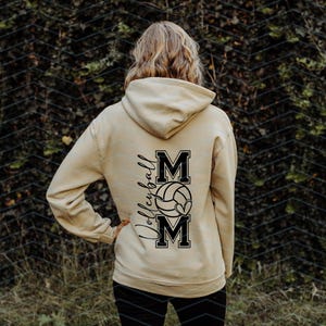 May include: A beige hoodie featuring the word "Volleyball" in script and the word "MOM" in block letters, with a volleyball graphic. The hoodie is worn by a person with blonde hair.