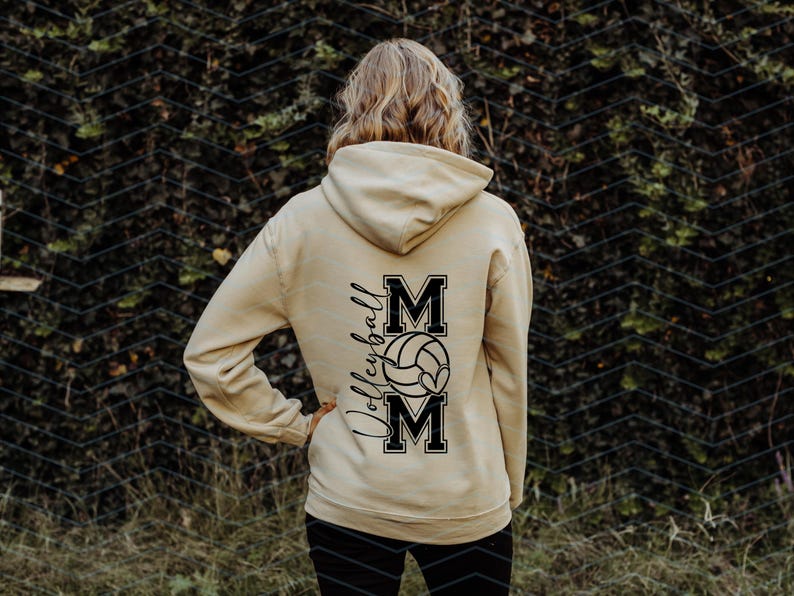 May include: A beige hoodie featuring the word "Volleyball" in script and the word "MOM" in block letters, with a volleyball graphic. The hoodie is worn by a person with blonde hair.