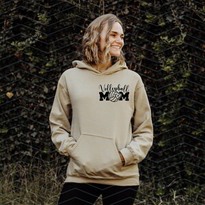 May include: A beige hoodie with a front pocket and a hood. The black text on the front says "Volleyball MOM" with a volleyball graphic. The person is standing in front of a green leafy background.