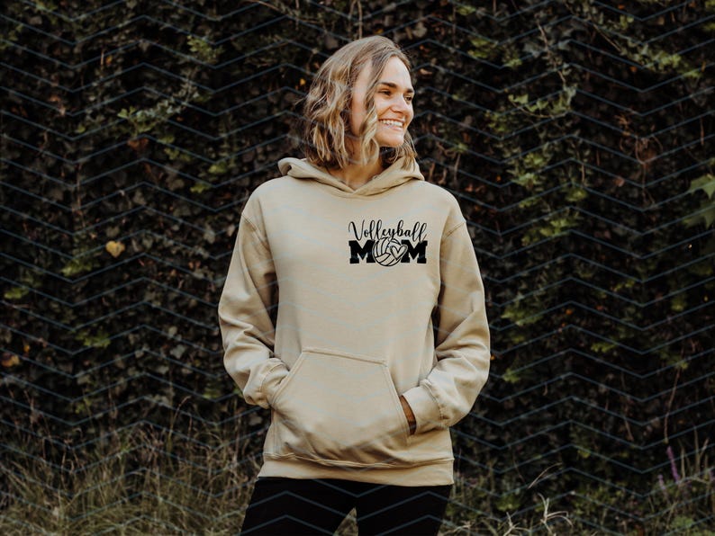 May include: A beige hoodie with a front pocket and a hood. The black text on the front says "Volleyball MOM" with a volleyball graphic. The person is standing in front of a green leafy background.