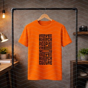 May include: An orange t-shirt with the text "Love Black People Like You Love Black Culture" in black, displayed on a black clothing rack. The shirt is hanging on a wooden hanger.