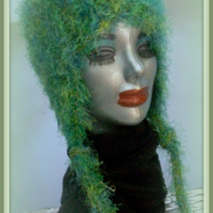 May include: A green and blue fuzzy hat with long strands of yarn hanging down the back. The hat is worn by a mannequin with a black turtleneck.