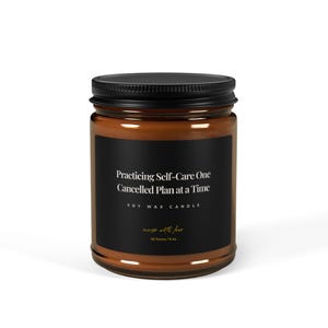 以下が含まれることがあります： 黒い蓋とラベルが付いた茶色のガラス製のキャンドルジャー。ラベルには「Practicing Self-Care One Cancelled Plan at a Time」と「Soy Wax Candle」と書かれています。